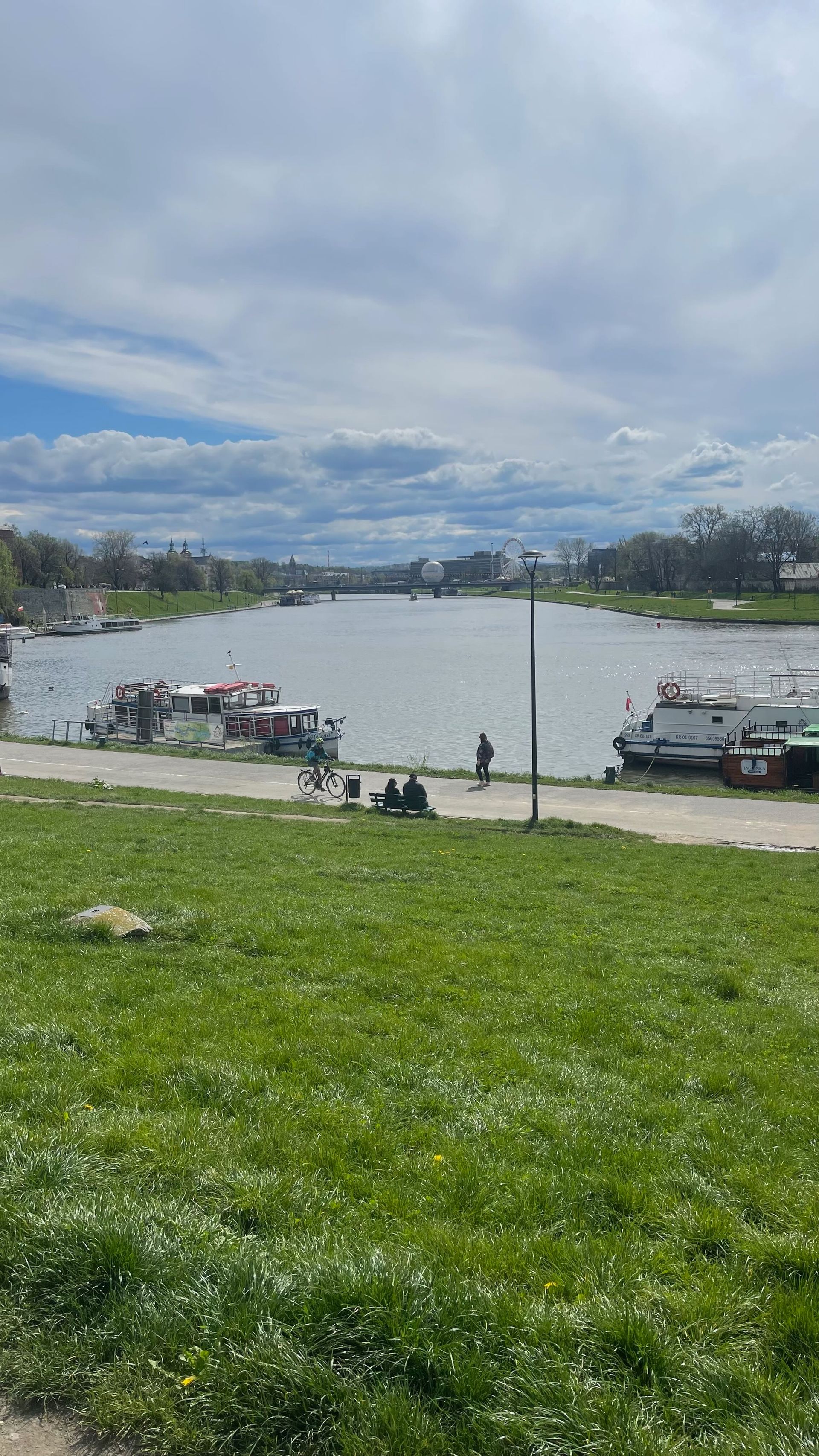 River scene with boats docked along the shore, green grass in the foreground, and cloudy sky.