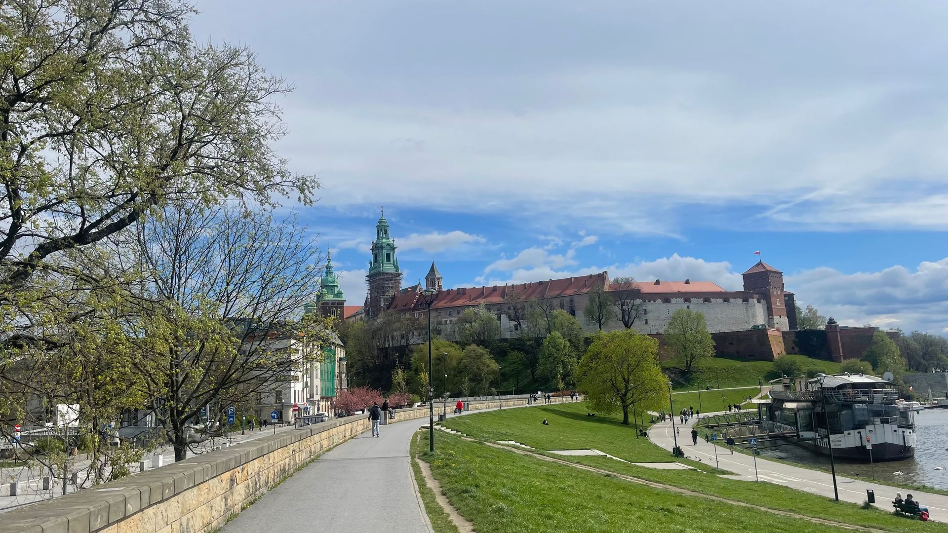 Wawel Castle in Krakow, Poland, seen from a riverside path. Blue sky, green grass, and trees are in the setting.