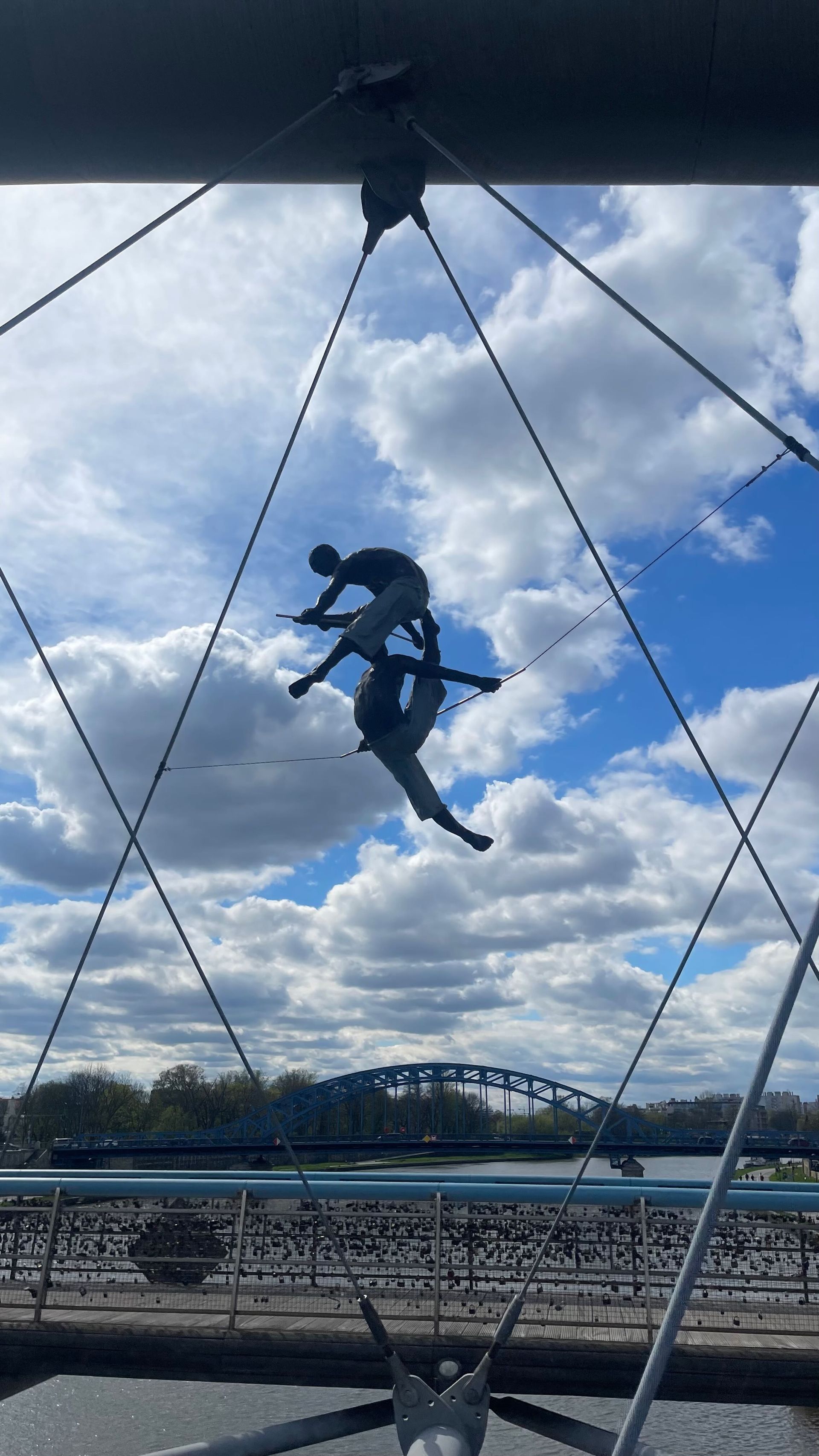 Sculpture of two figures suspended in the air against a bridge and cloudy sky backdrop.