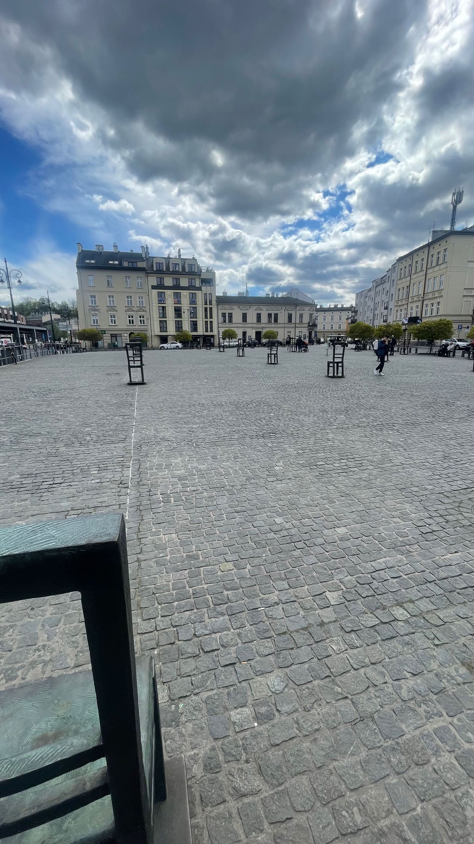 Cobblestone square with buildings under a cloudy sky. People are walking.