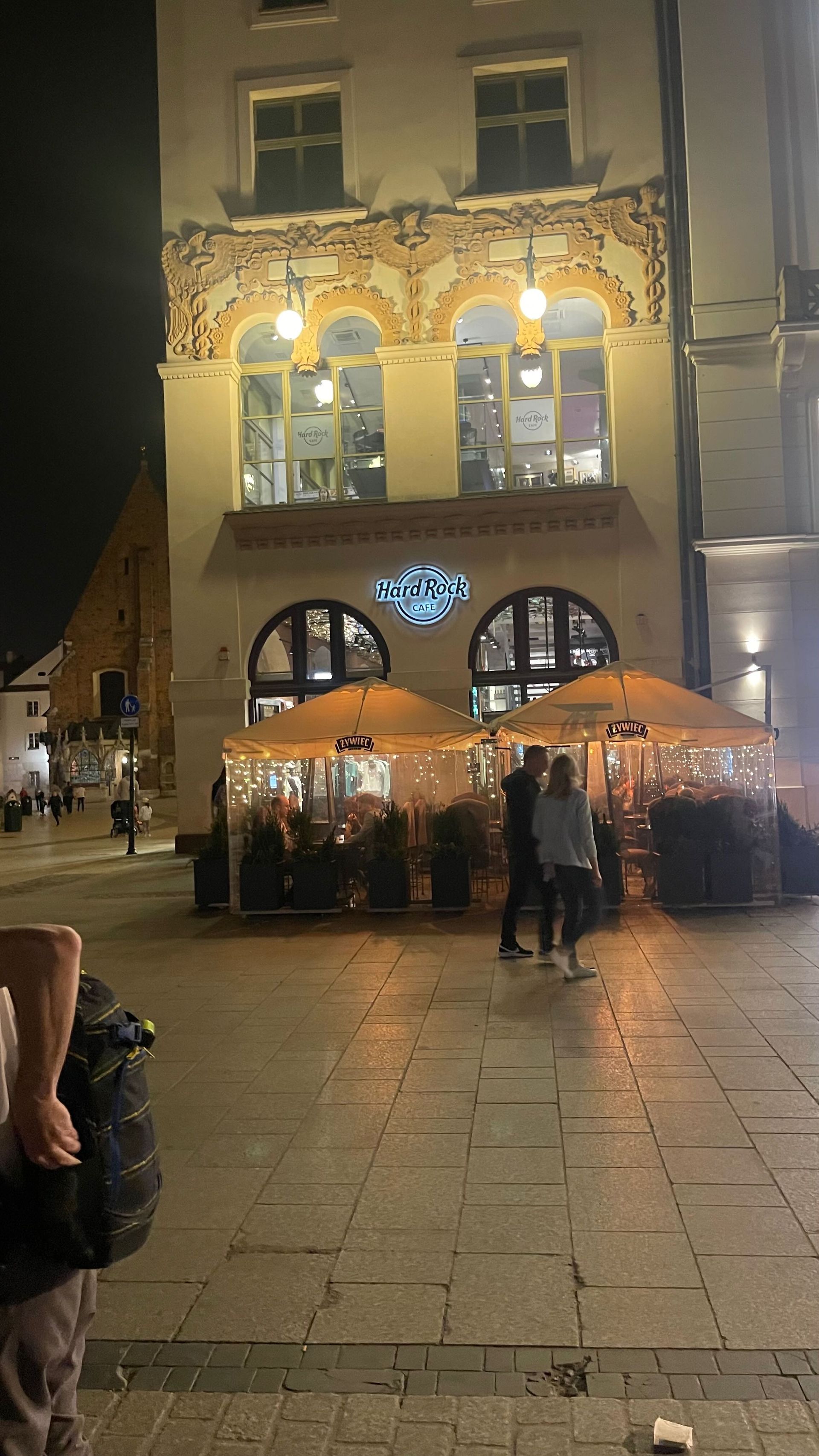 Restaurant with outdoor seating at night, people walking, building with ornate architecture, street.