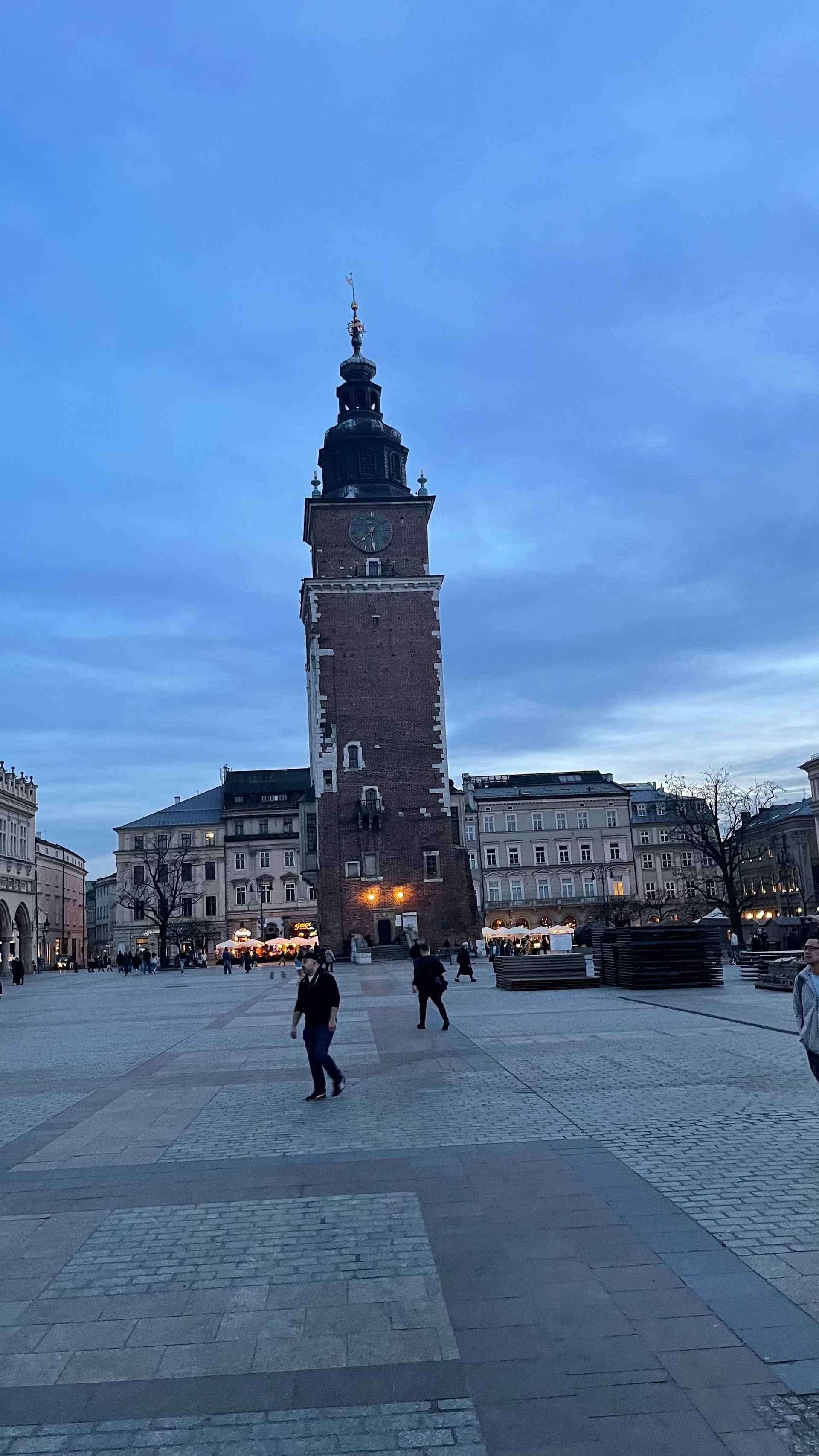 Brick tower in a cobblestone square; people walk by; blue sky overhead; Krakow, Poland.