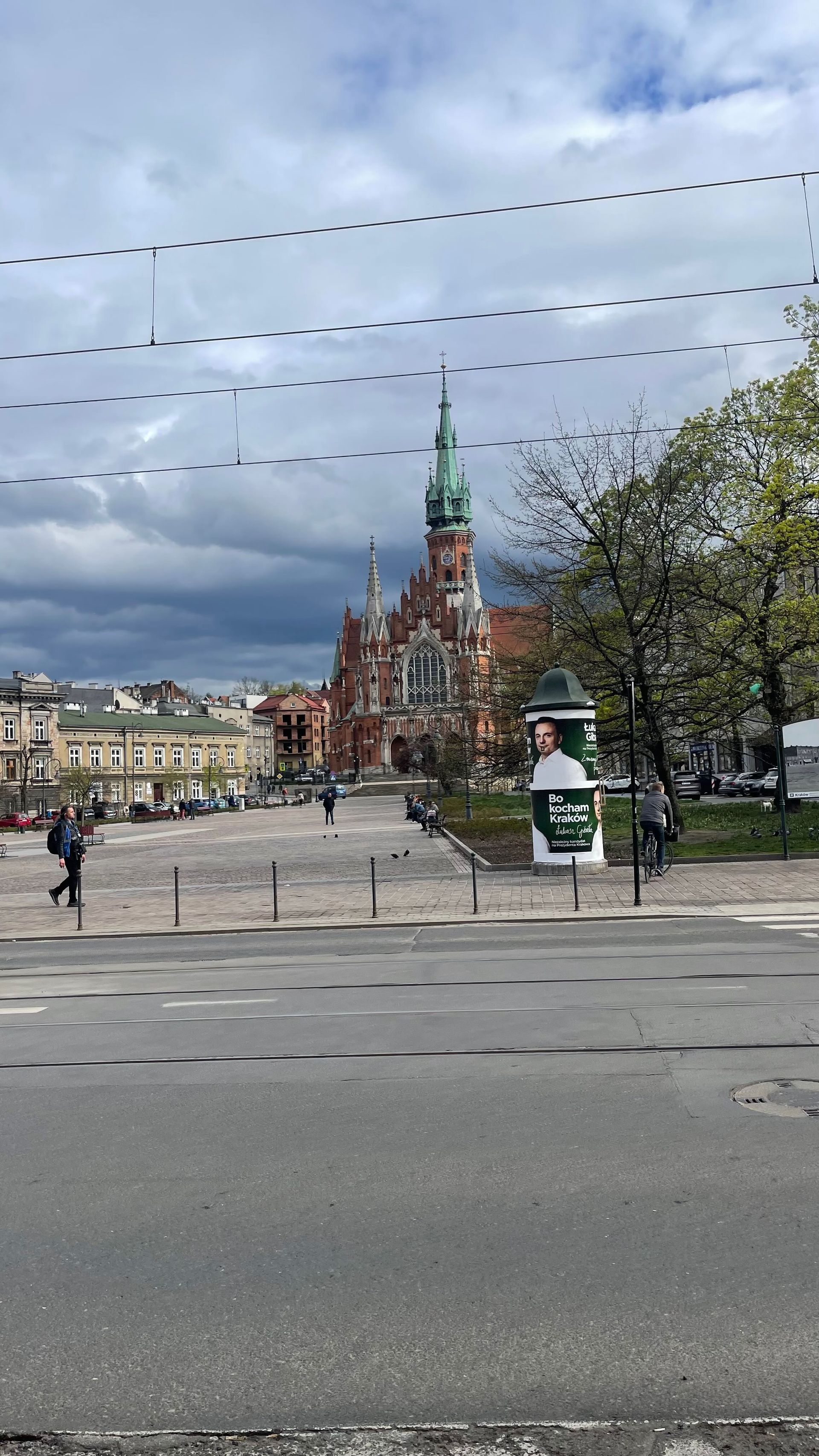 Church with a tall green spire in a city square, cloudy sky above. People walking and a street in front.