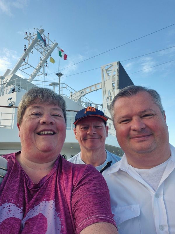 Three people smiling in front of a cruise ship. Blue sky, sunny day.