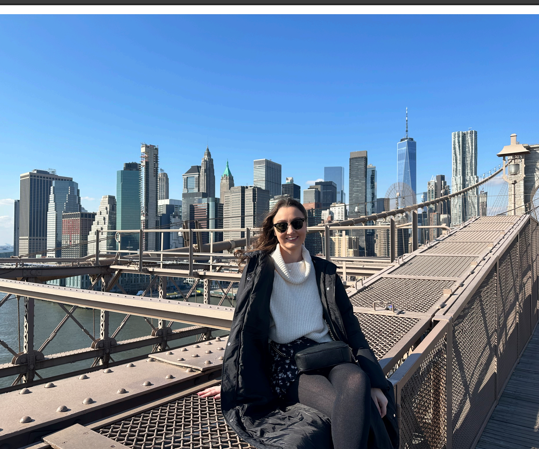 Woman sitting on the Brooklyn Bridge with Manhattan skyline in the background. She is smiling.