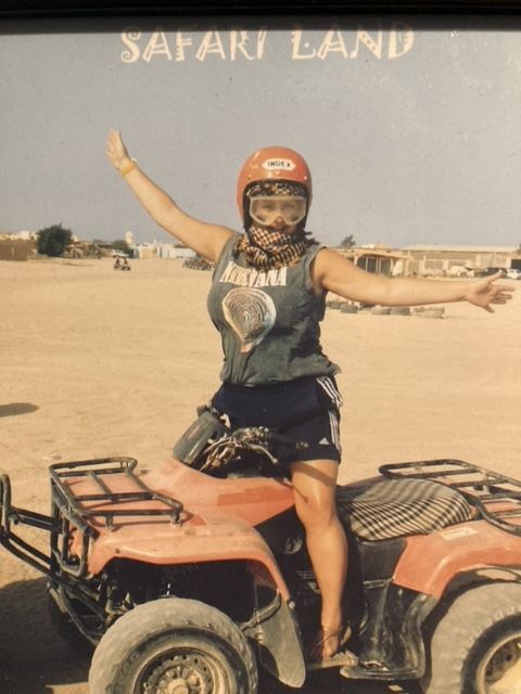 Woman on an ATV with arms outstretched, wearing helmet, sunglasses, and dust protector, in desert terrain.