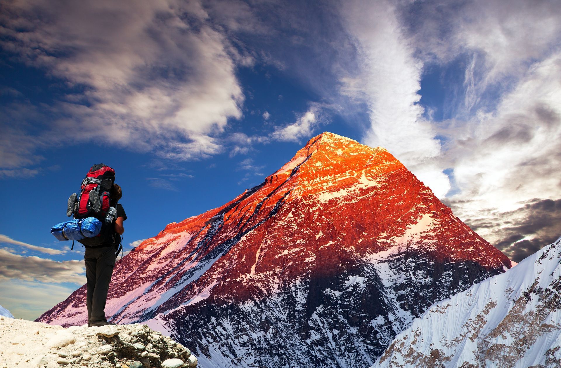 Person with backpack overlooking a snow-covered mountain at sunset, blue sky with clouds.