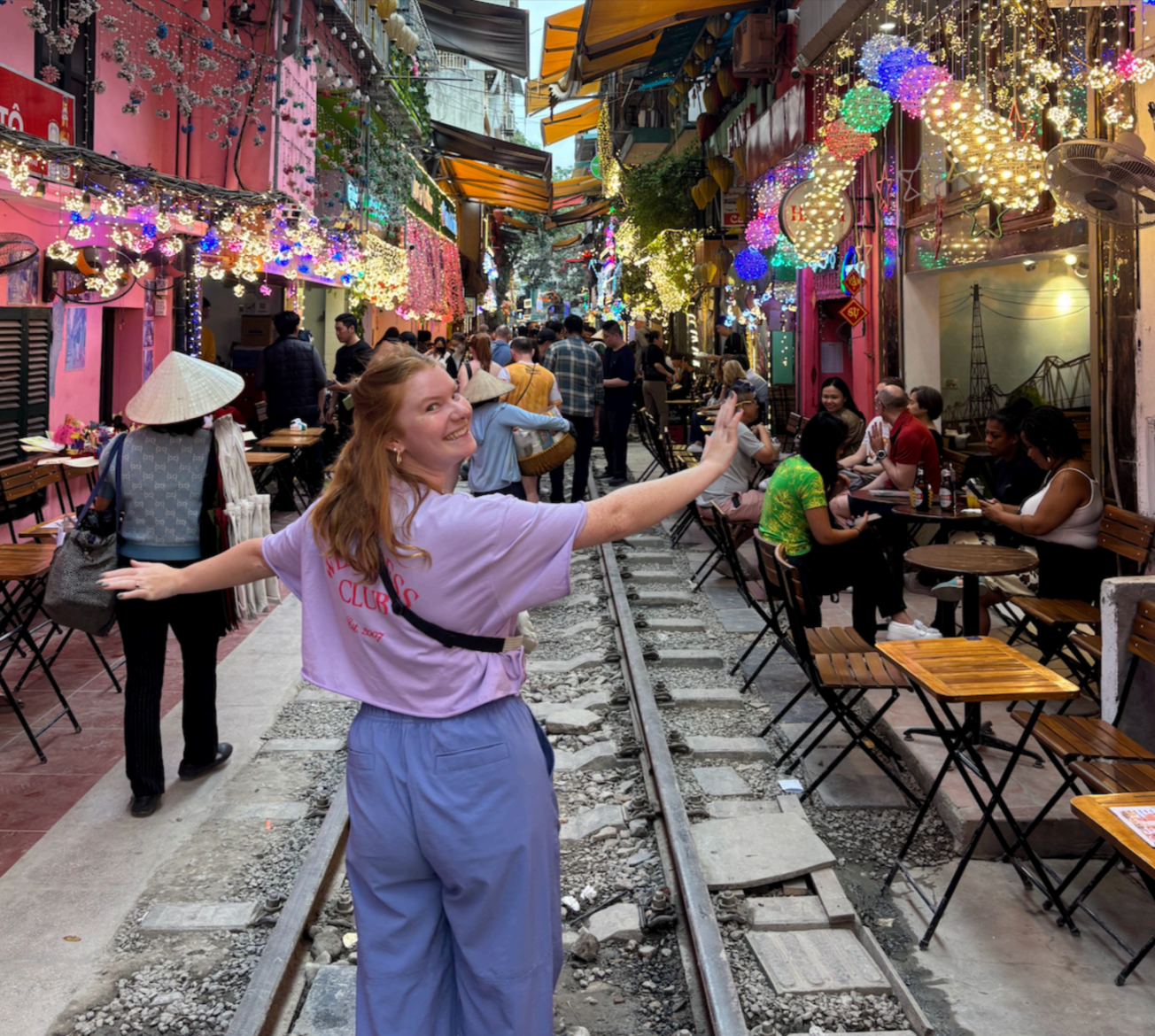 Woman with arms outstretched smiles on train tracks in a colorful alley, tables, and string lights.