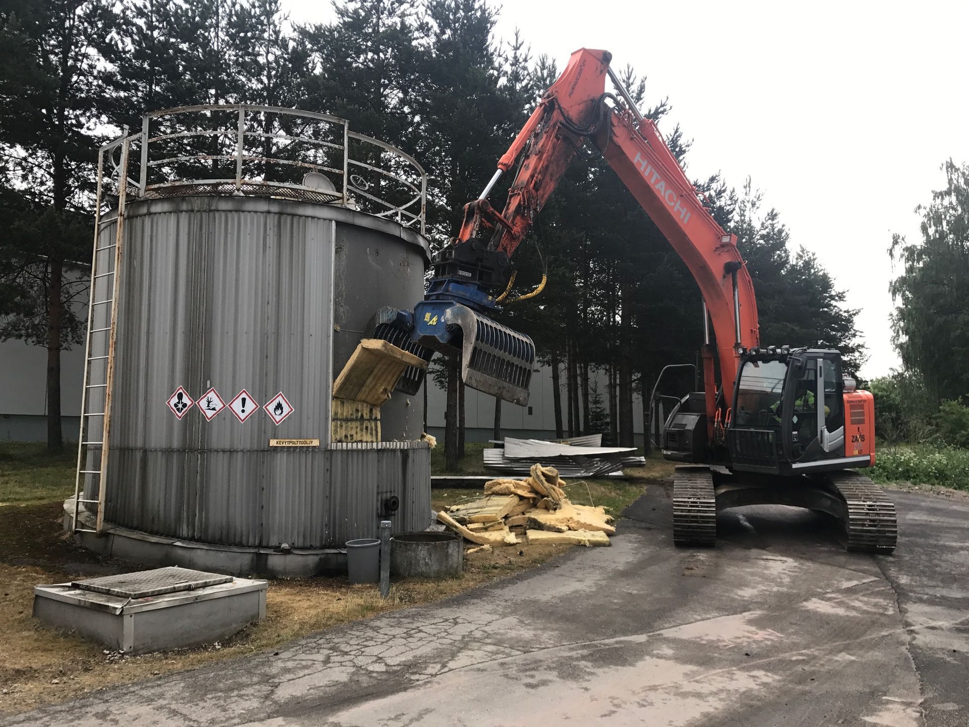 Oranssi kaivinkone käyttää hydraulista kouraa metallista teollisuussäiliötä repimiseen ulkotontilla.
