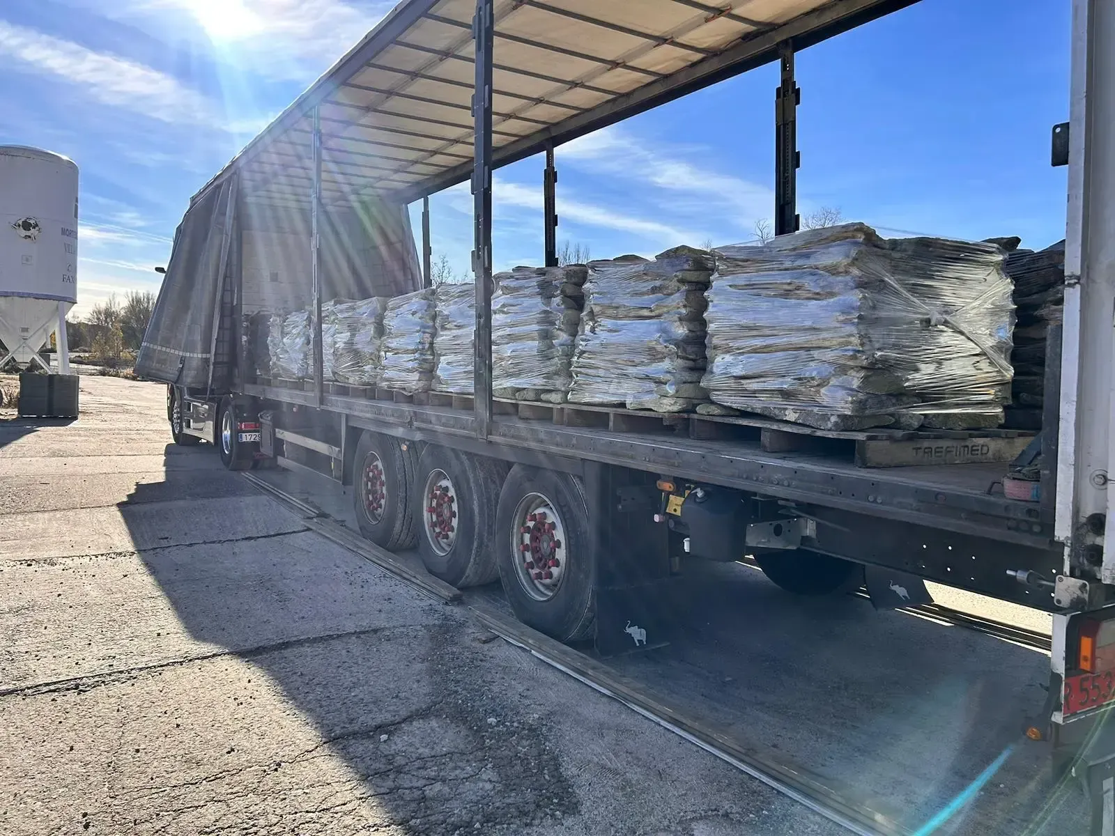 Remolque de plataforma cargado con materiales apilados y envueltos en papel blanco en un patio industrial.