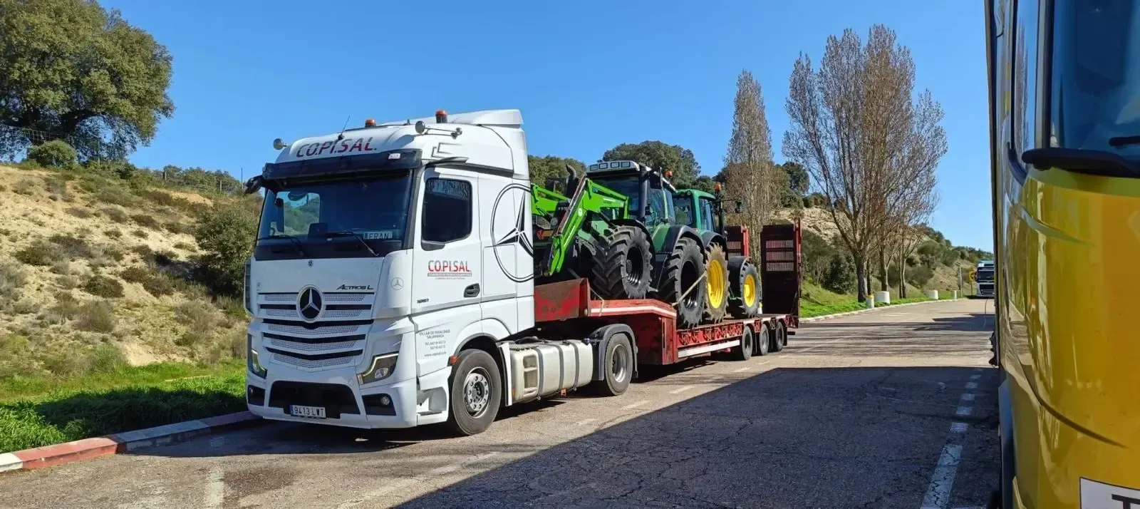 Camión Mercedes blanco transportando maquinaria agrícola verde en una carretera rural bajo un cielo azul.