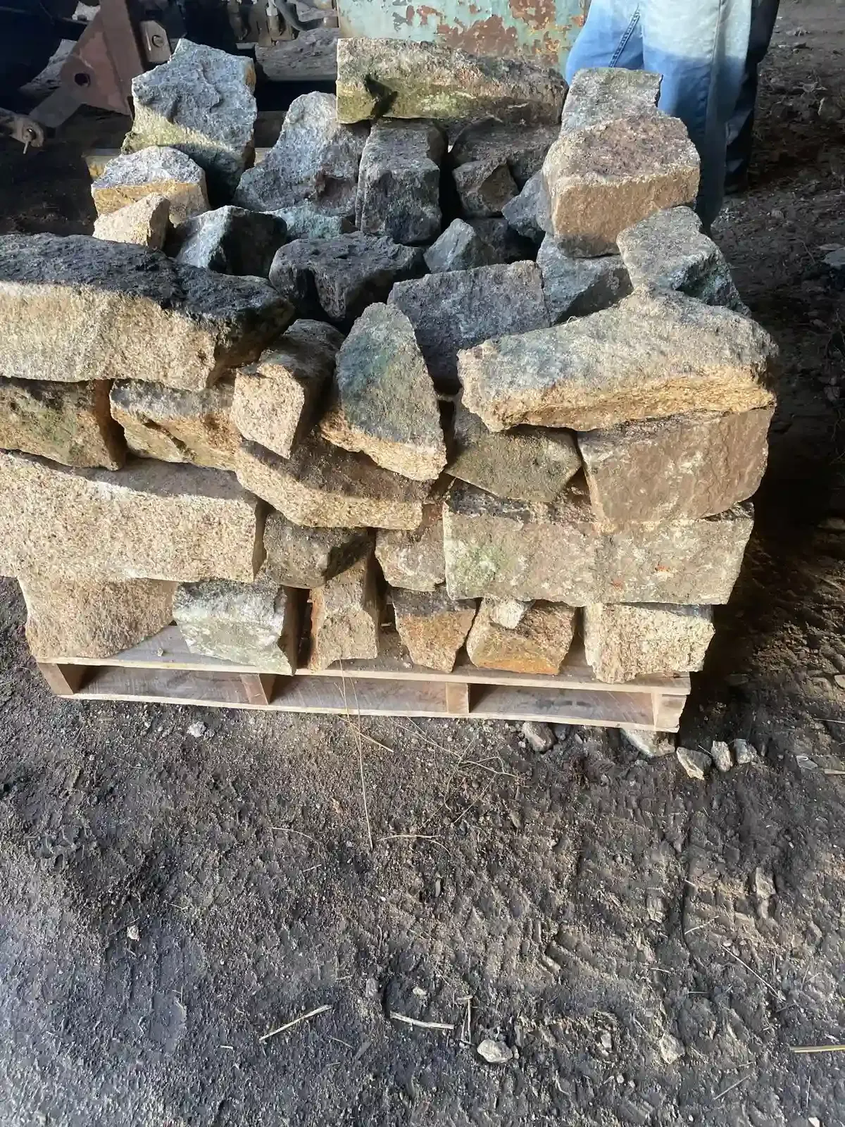 Piedras toscas apiladas sobre un palé de madera al aire libre, sobre suelo de tierra.