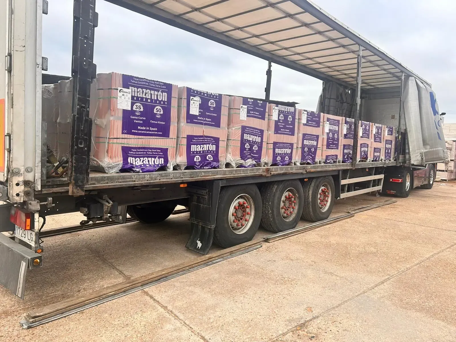 Semirremolque cargado con cajas apiladas de color morado y blanco, estacionado en el muelle de un almacén.