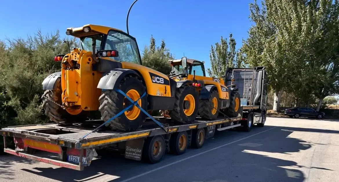 Máquina de construcción naranja cargada en un remolque de plataforma estacionada junto a una carretera bajo un cielo azul despejado
