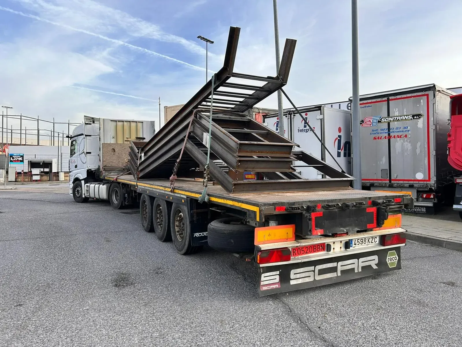 Camión de plataforma que transporta vigas metálicas apiladas en un patio industrial.