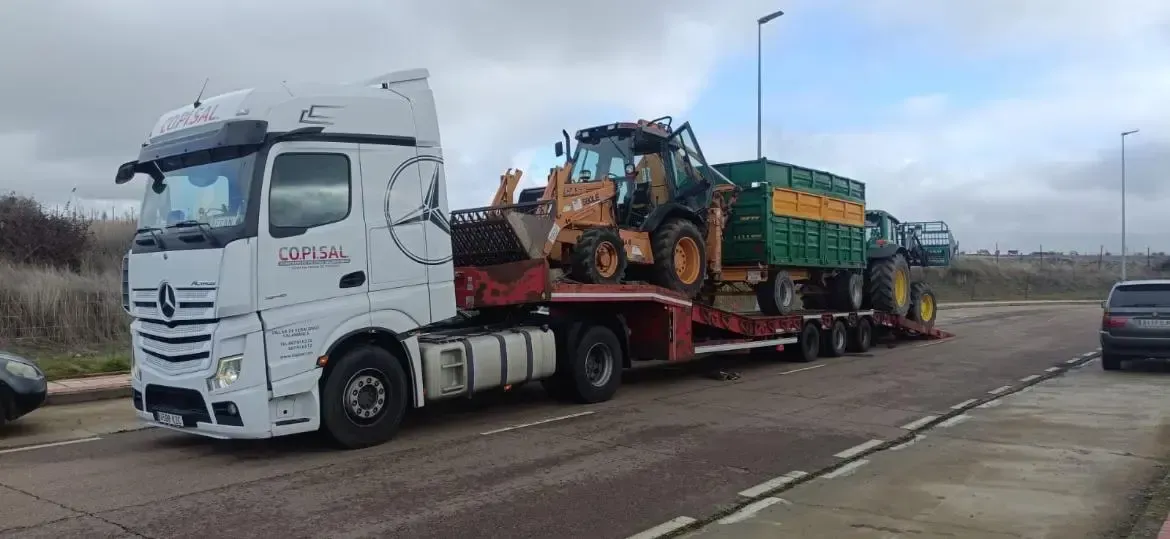 Camión Mercedes blanco transportando una cargadora en un remolque de plataforma roja en el arcén de una autopista.