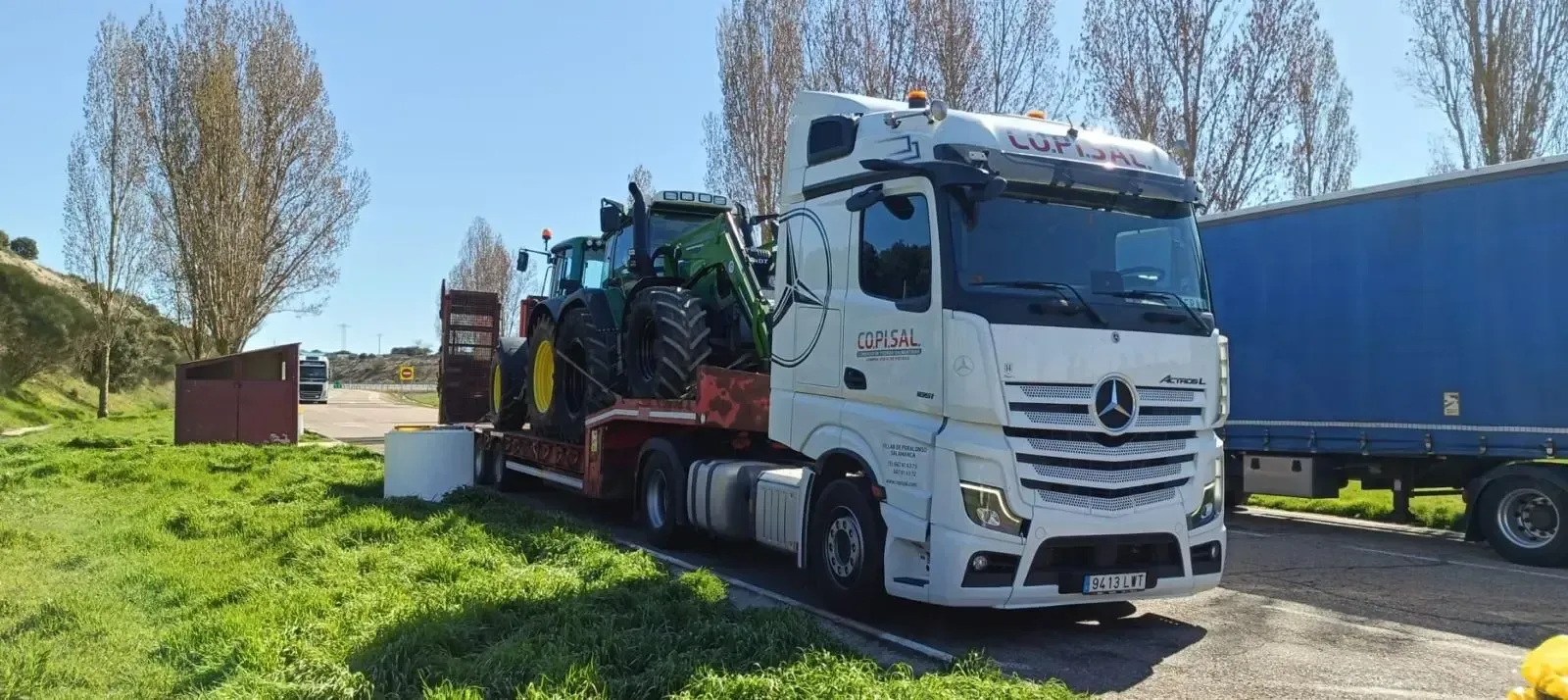 Un camión Mercedes blanco cargado de maquinaria agrícola estacionado junto a un remolque azul en una carretera soleada.