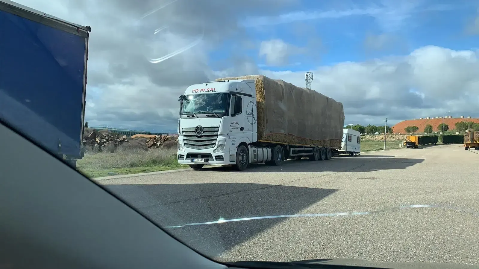 Camión semirremolque blanco con remolque cubierto estacionado al borde de la carretera bajo un cielo nublado, visto desde la ventana de un coche.