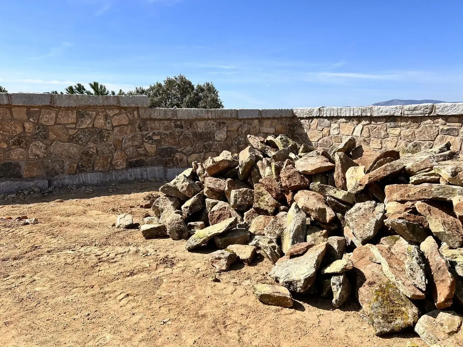 Montón de rocas y escombros junto a un muro bajo de piedra bajo un cielo azul despejado.
