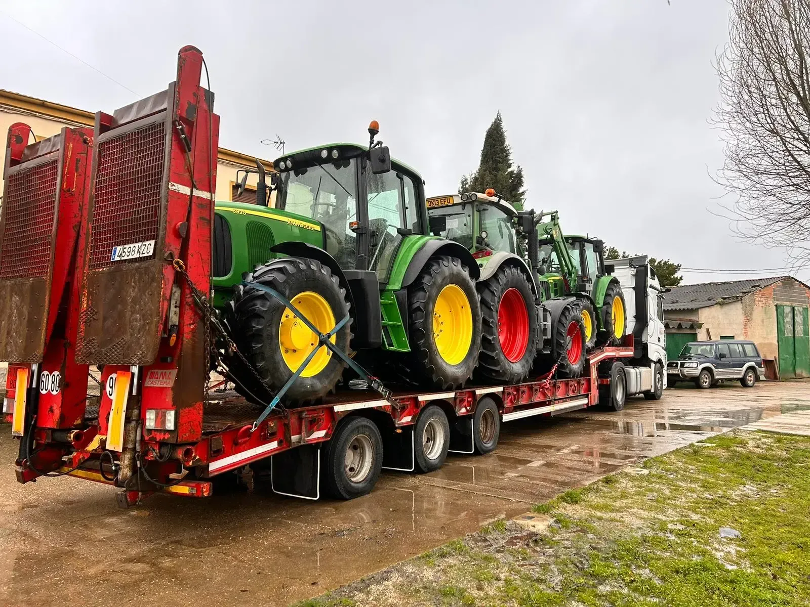 Un camión con remolque rojo que transporta varios tractores verdes en una carretera mojada cerca de edificios.