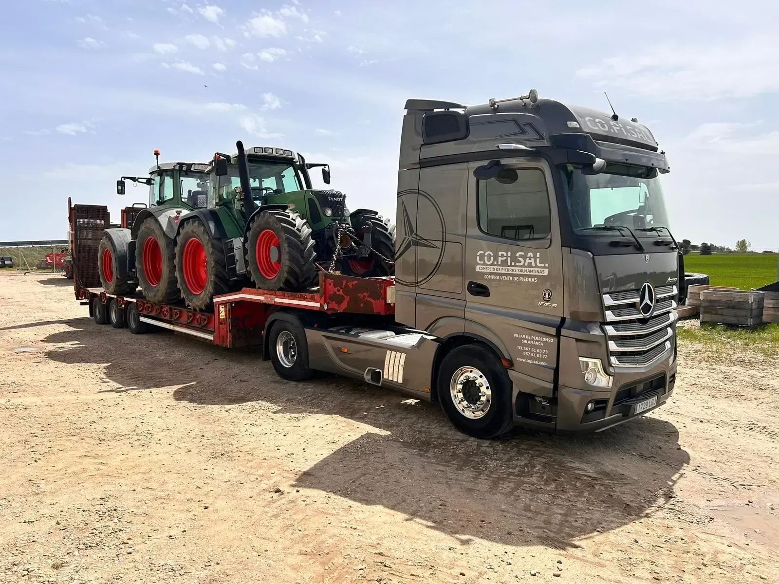 Un camión con remolque gris transporta dos tractores verdes en una plataforma en un campo rural.
