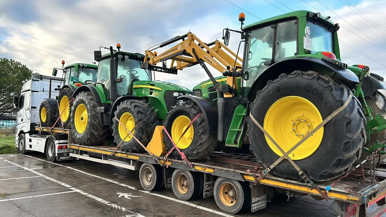 Dos tractores verdes con ruedas amarillas sujetos a un remolque de plataforma, vistos de lado.