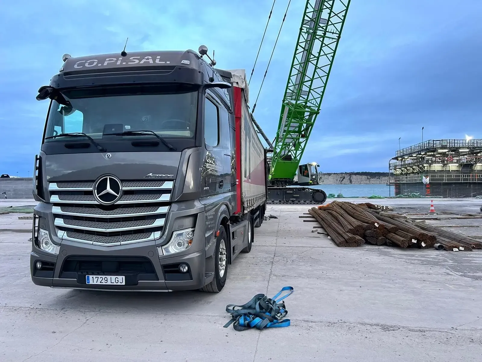 Un camión Mercedes gris estacionado en un puerto, con una grúa verde y troncos apilados al fondo.