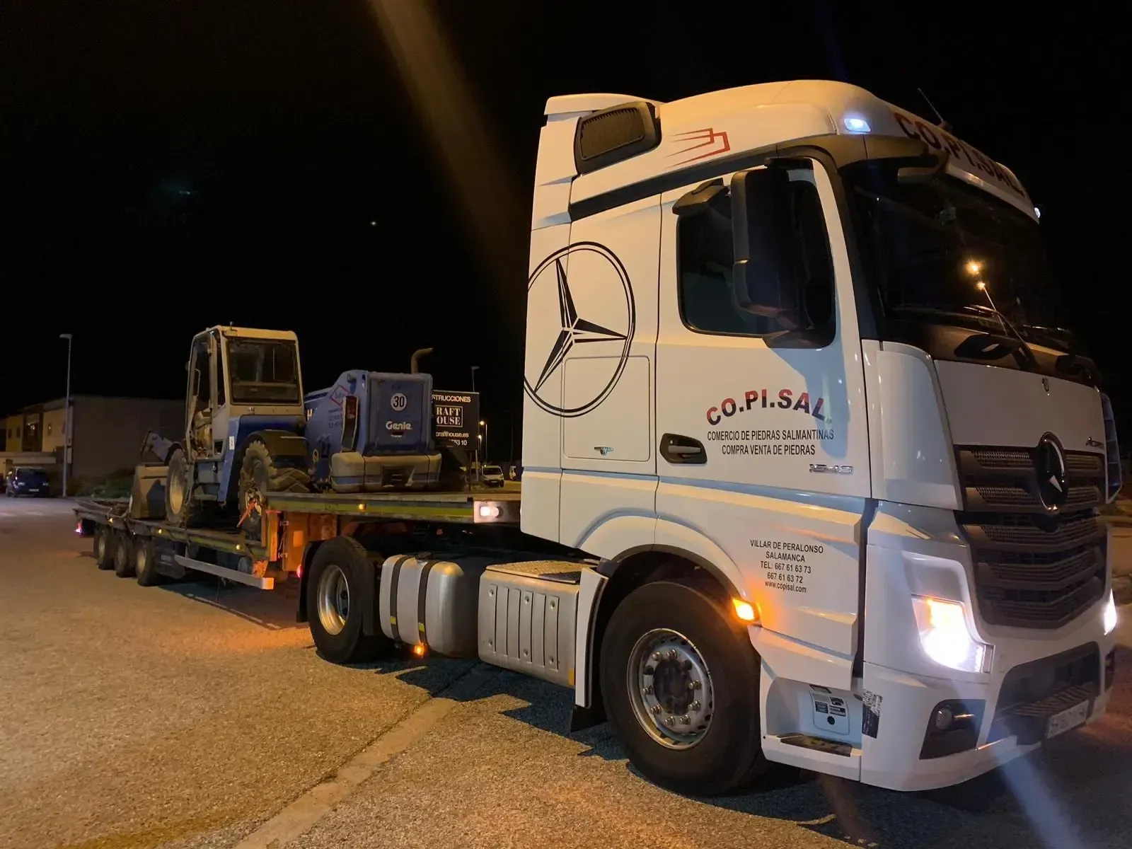 Camión blanco remolcando un remolque de plataforma con un segundo camión por la noche en una carretera.