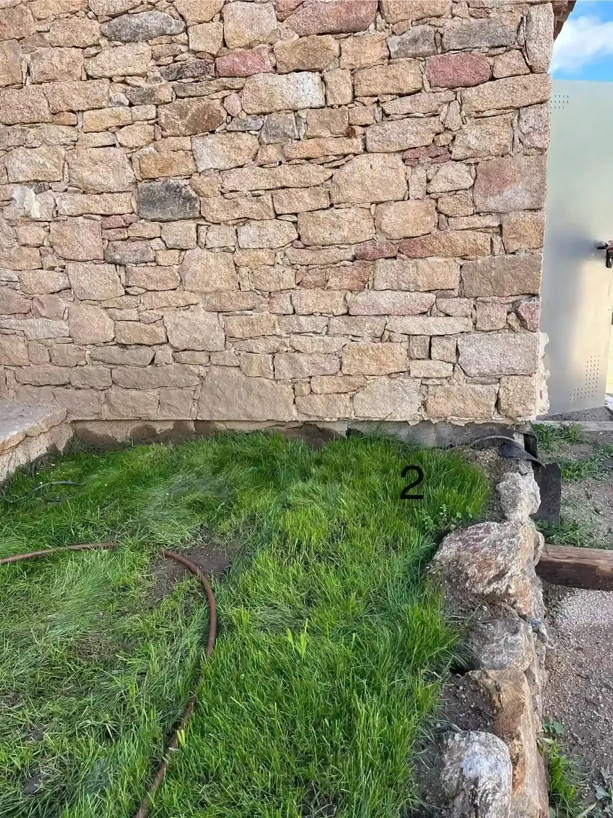 Muro de piedra junto a un pequeño patio con césped, un estrecho borde de hormigón y una vista parcial del cielo azul.