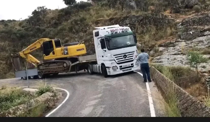 Un camión blanco que transportaba una excavadora amarilla quedó atascado en una carretera estrecha y sinuosa junto a una cuneta.