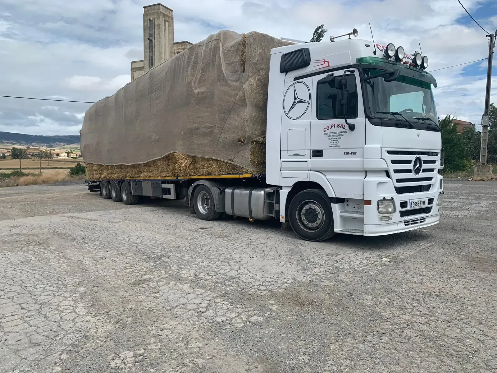 Camión Mercedes blanco transportando una enorme carga cubierta de rocas en un camino de grava en una zona rural.