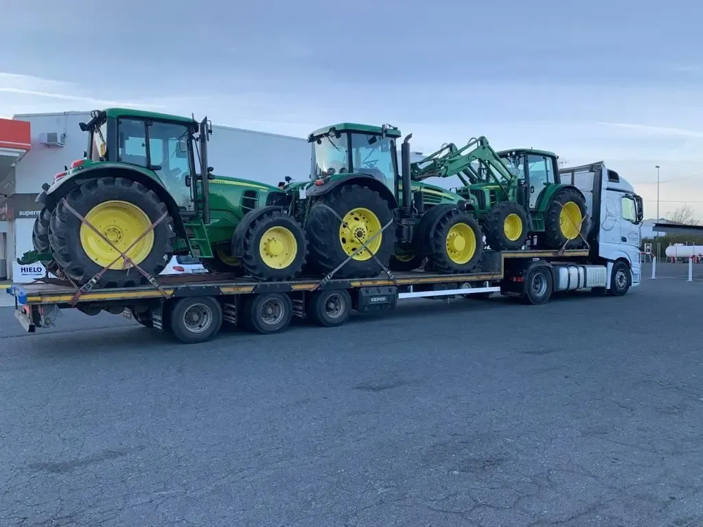 Tres tractores verdes con ruedas amarillas cargados en un remolque de plataforma junto a un camión semirremolque blanco.
