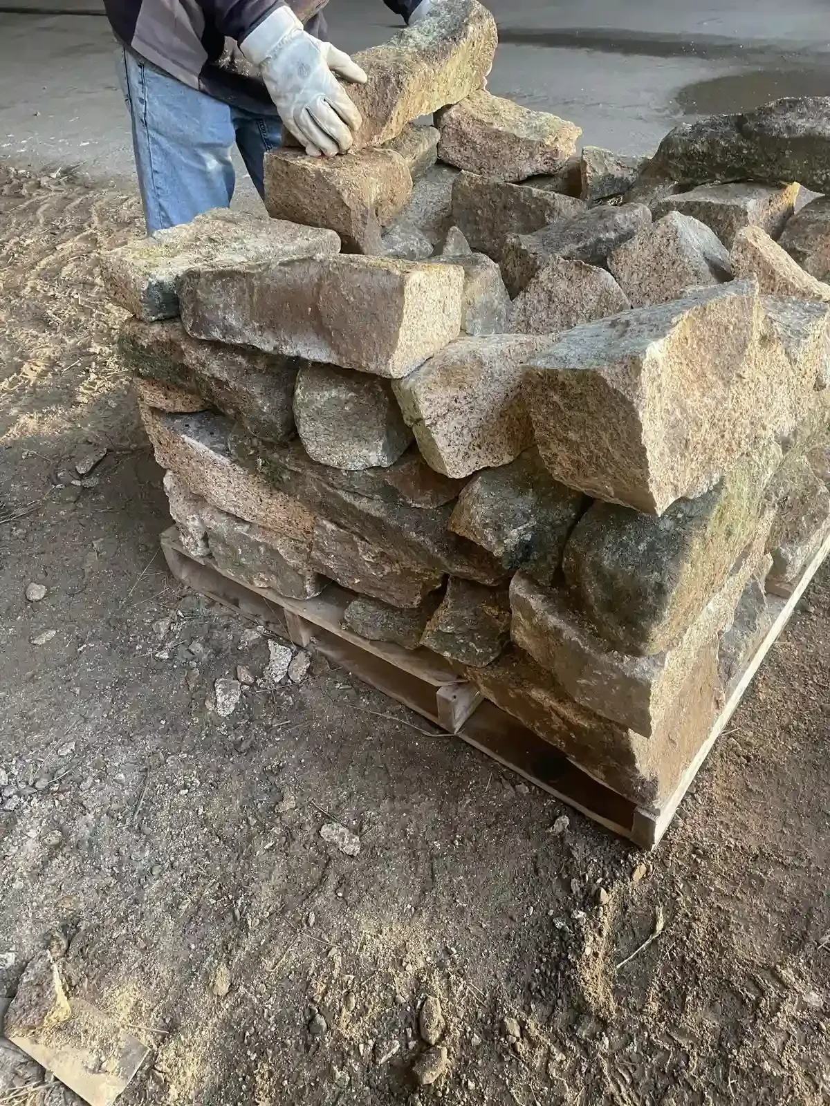 Pila de piedras toscamente labradas sobre un palé, colocadas al aire libre sobre una superficie de tierra por manos enguantadas.