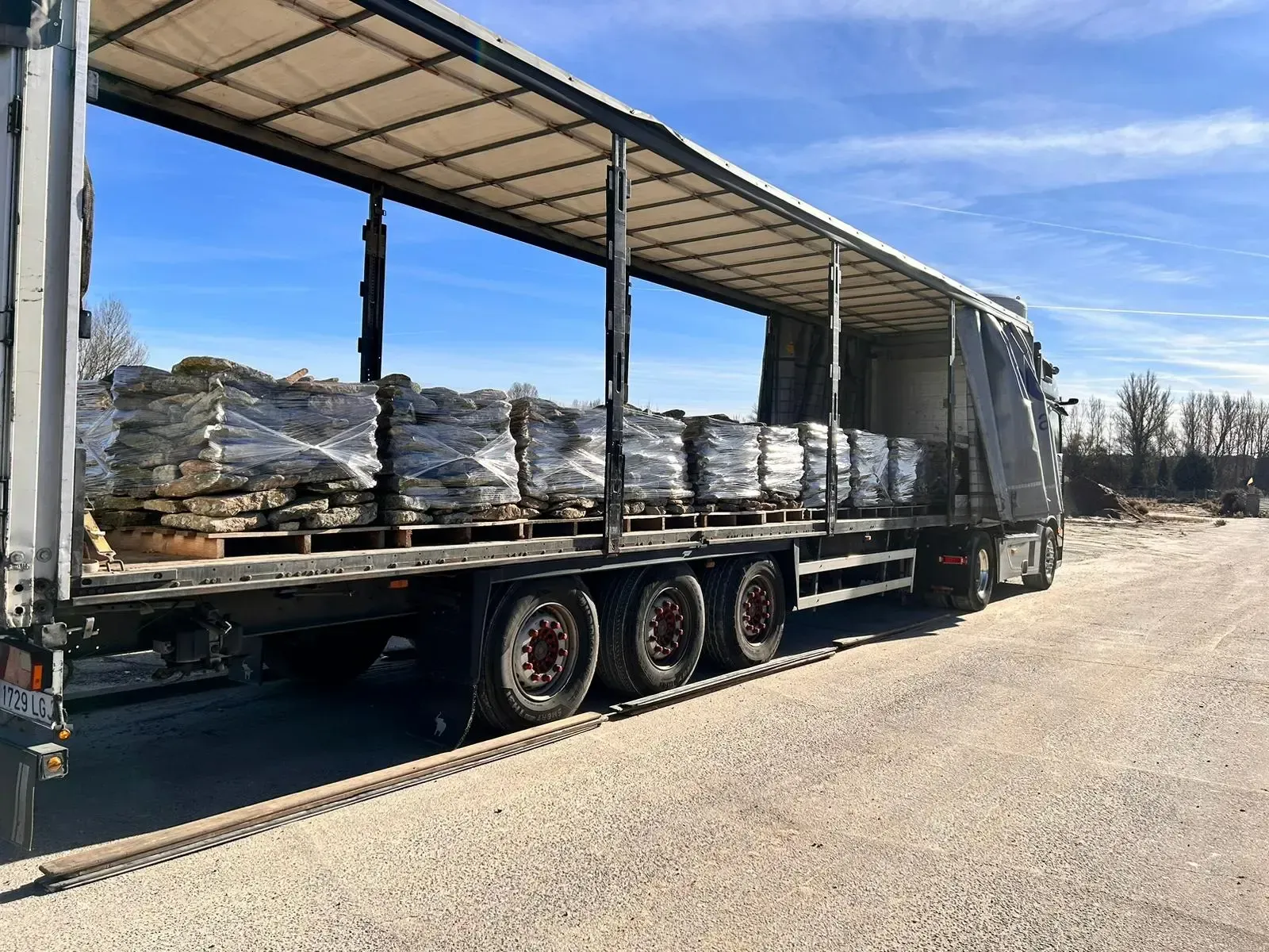 Camión de plataforma cargado con palés envueltos estacionado en un camino de tierra bajo un cielo azul despejado.