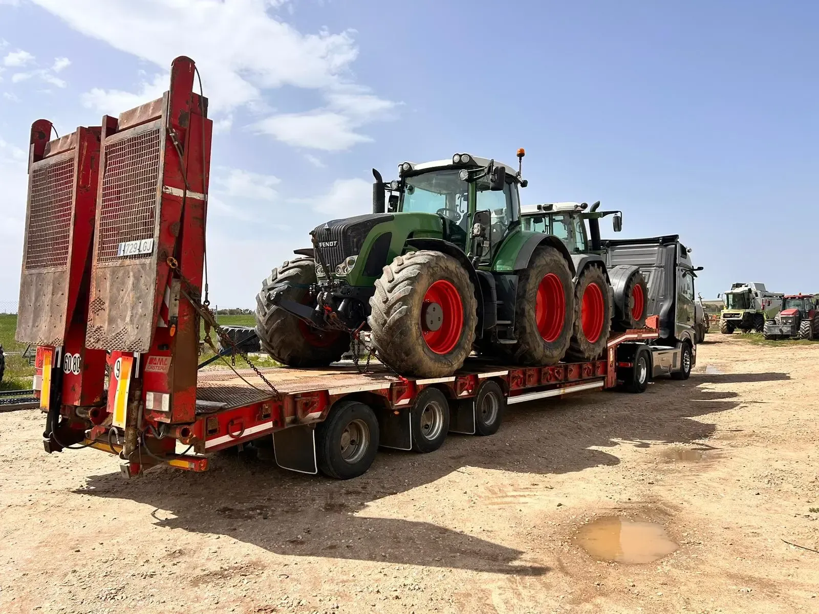 Un remolque de plataforma roja transporta un gran tractor verde en un terreno de tierra, con otro tractor al fondo.