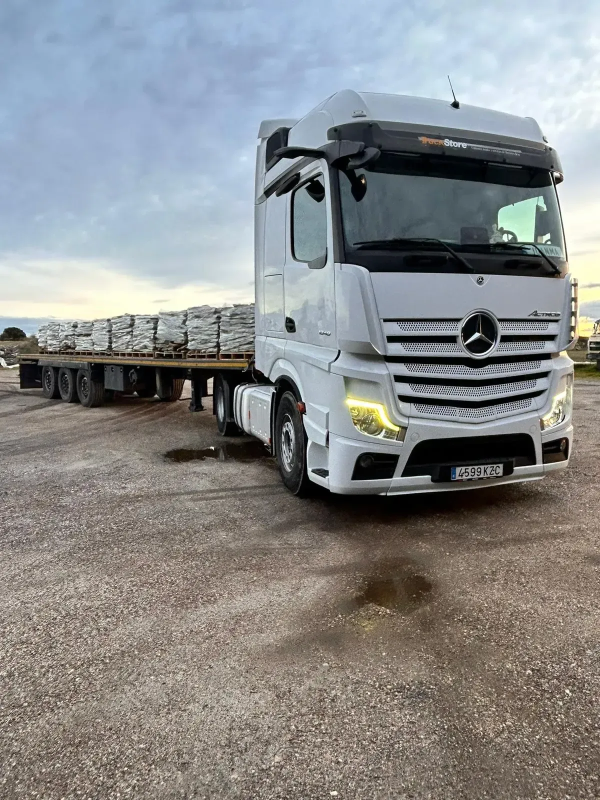 Camión semirremolque blanco transportando un remolque de plataforma cargado de materiales en un terreno de grava.