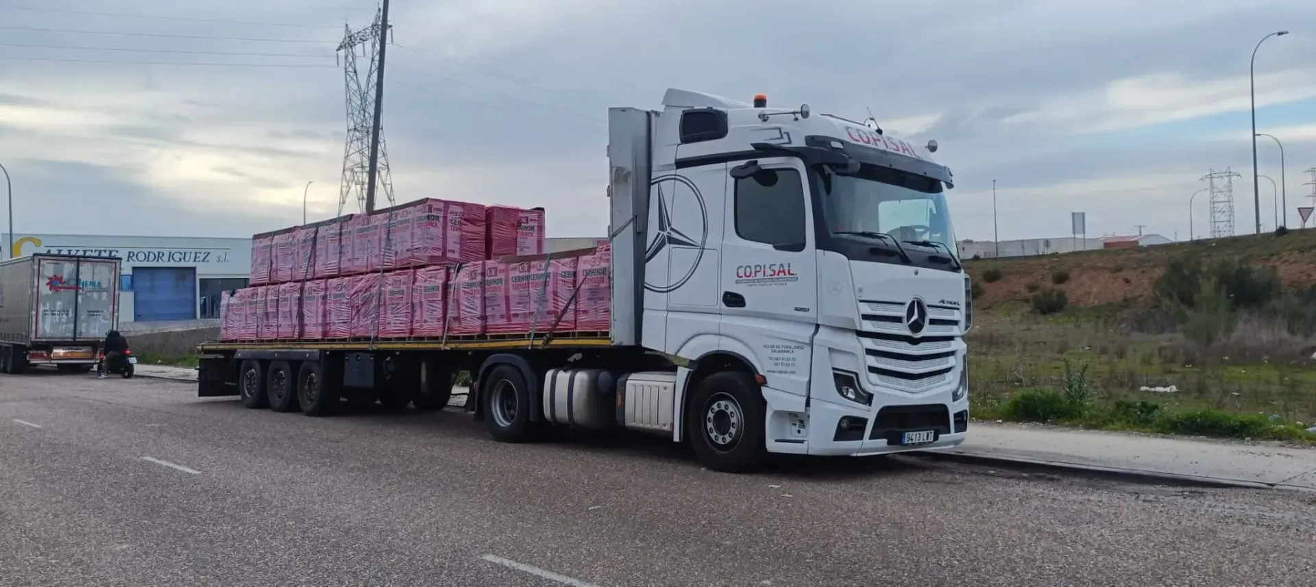 Un camión semirremolque blanco transporta carga envuelta en rosa por una autopista bajo un cielo nublado.