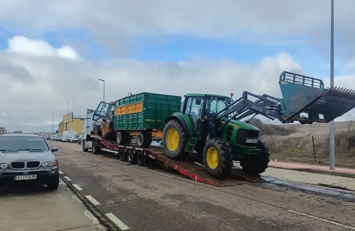 Un tractor verde carga un remolque agrícola en un camión de plataforma en la cuneta, bajo un cielo nublado.