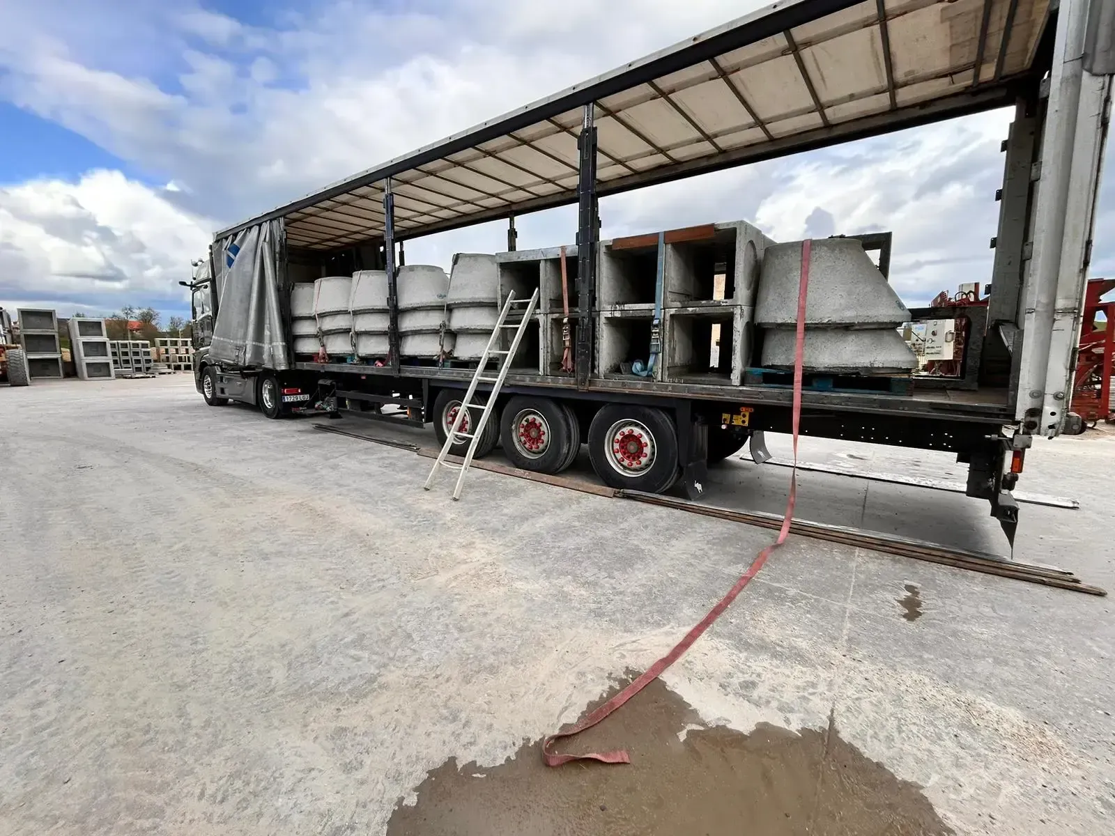 Camión de plataforma que transporta grandes tuberías de hormigón bajo un toldo en un terreno de grava, con una escalera al lado.