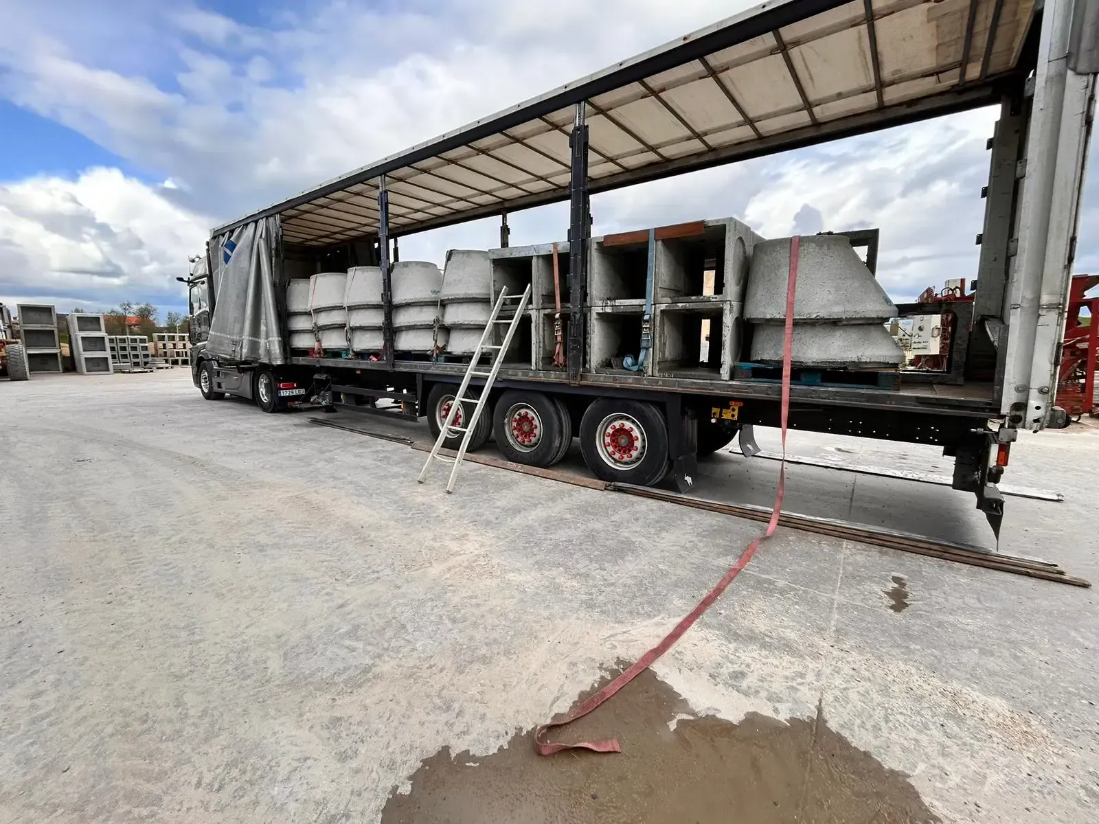 Camión de plataforma cargado con barreras de hormigón bajo un toldo en un terreno de grava, con una correa roja en el suelo.