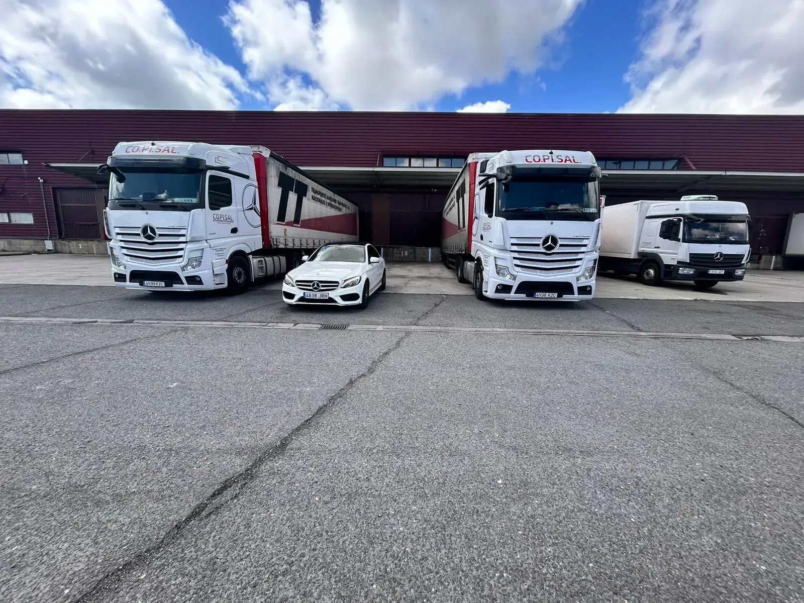 Camiones blancos y un pequeño coche blanco estacionados frente a un almacén bajo un cielo nublado.