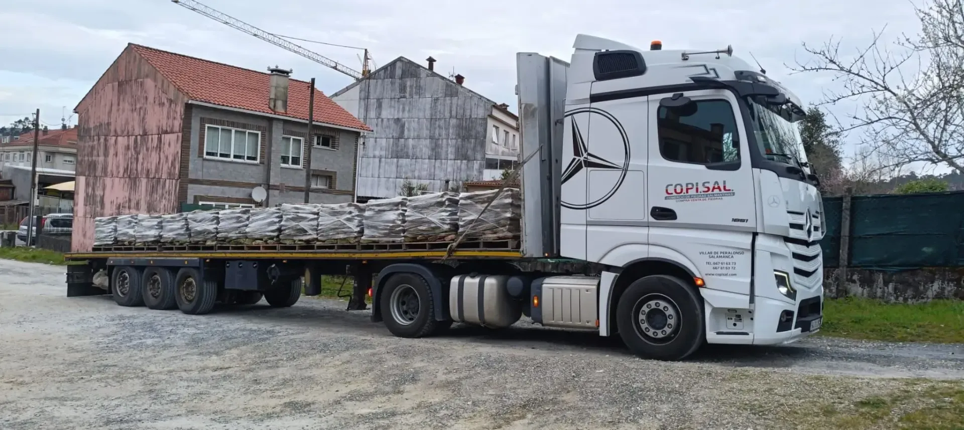 Un camión semirremolque blanco transporta una casa rosa dañada en un remolque de plataforma en un terreno de grava.