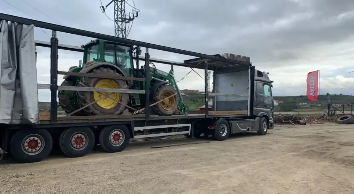 Un gran camión con remolque transporta un tractor verde con chasis en un terreno de grava bajo un cielo nublado.