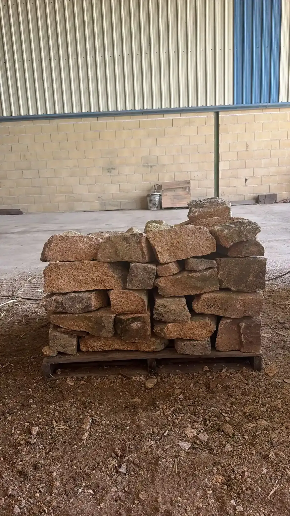 Ladrillos apilados sobre un palé en un almacén o patio, contra una pared de chapa ondulada.