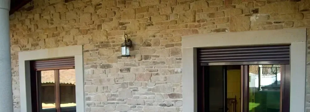 Muro exterior de piedra con dos ventanas y una lámpara de pared entre ellas.