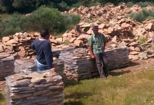 Dos personas están de pie junto a unos muros de piedra apilados en una zona cubierta de hierba, con un montón de rocas detrás de ellas.