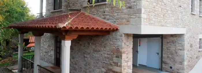 Exterior de una casa de piedra con un pequeño porche cubierto de tejas rojas y una entrada con escalera.