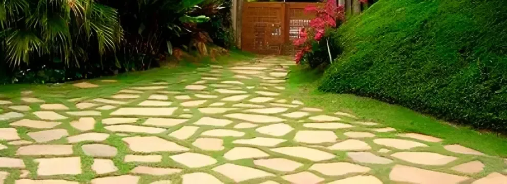 Un sendero de piedra atraviesa un jardín verde exuberante que conduce a una puerta de madera y flores rojas.