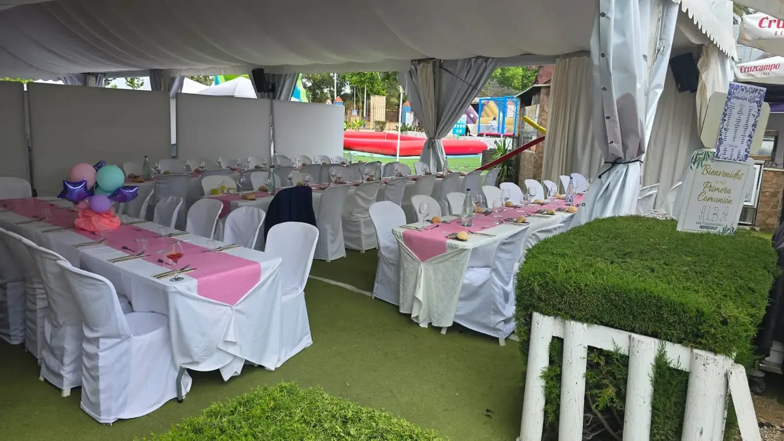 Una carpa blanca con largas mesas de banquete, sillas blancas, alfombras rosas y decoraciones con globos sobre un césped.