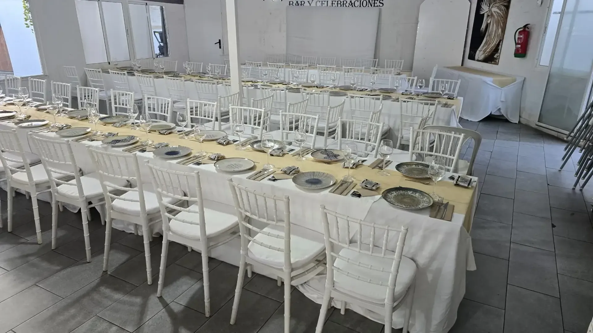 Mesa larga para banquetes con sillas blancas, cristalería y bajoplatos dorados en un luminoso salón de eventos.